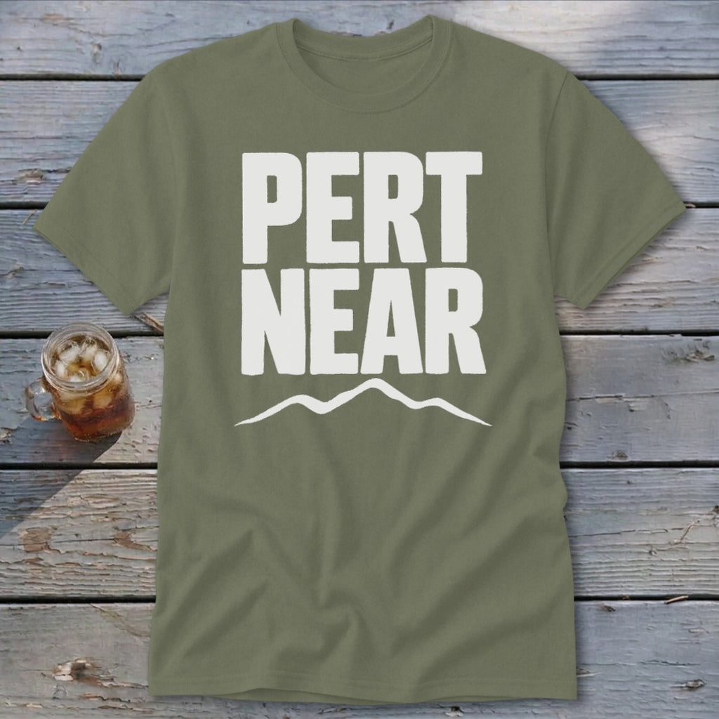 An olive green t-shirt with white text reading PERT NEAR above a mountain outline rests on weathered wooden boards beside a glass of iced tea.