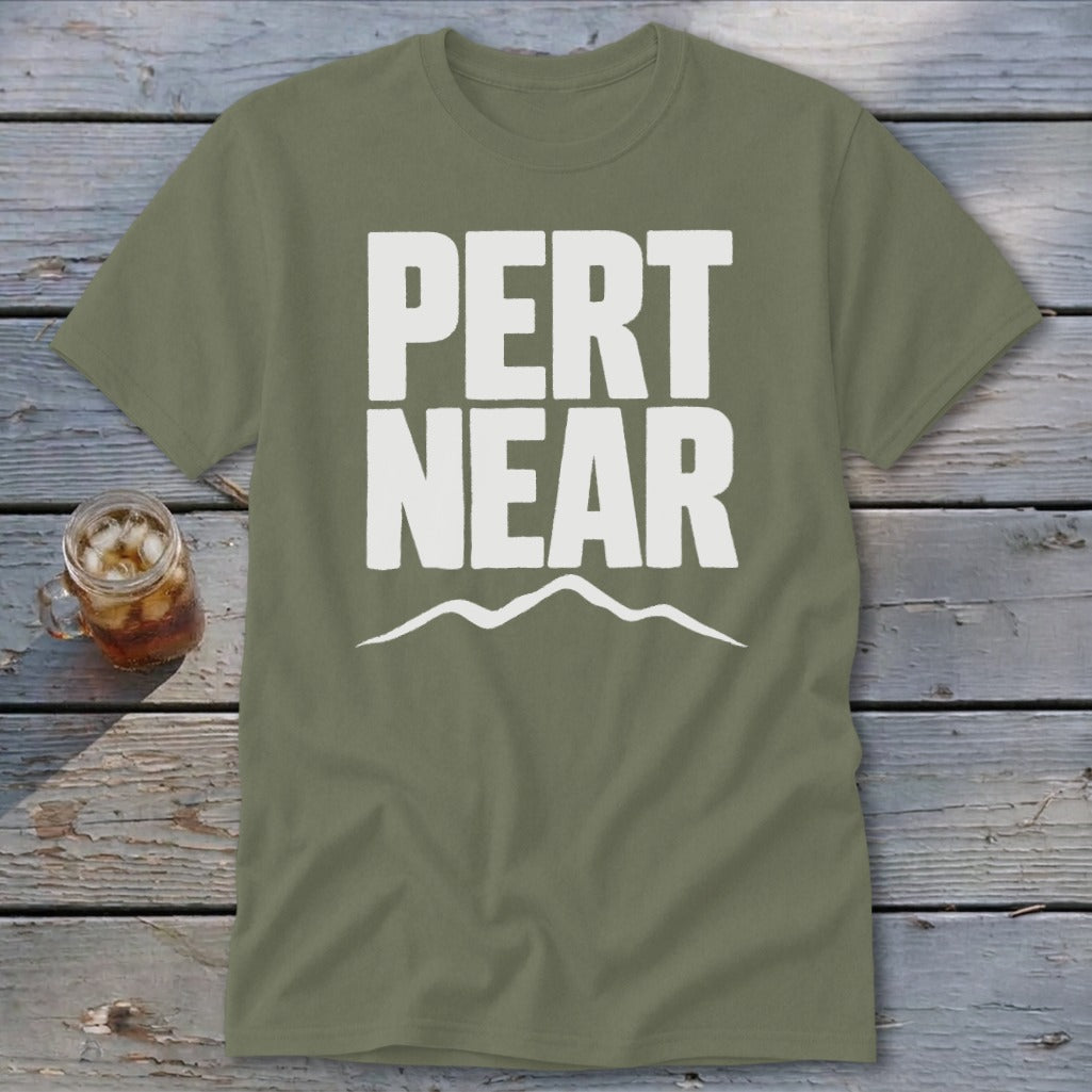 An olive green t-shirt with white text reading PERT NEAR above a mountain outline rests on weathered wooden boards beside a glass of iced tea.