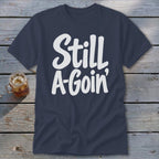 Dark blue t-shirt featuring the white text Still A-Goin' laid flat on wooden planks.