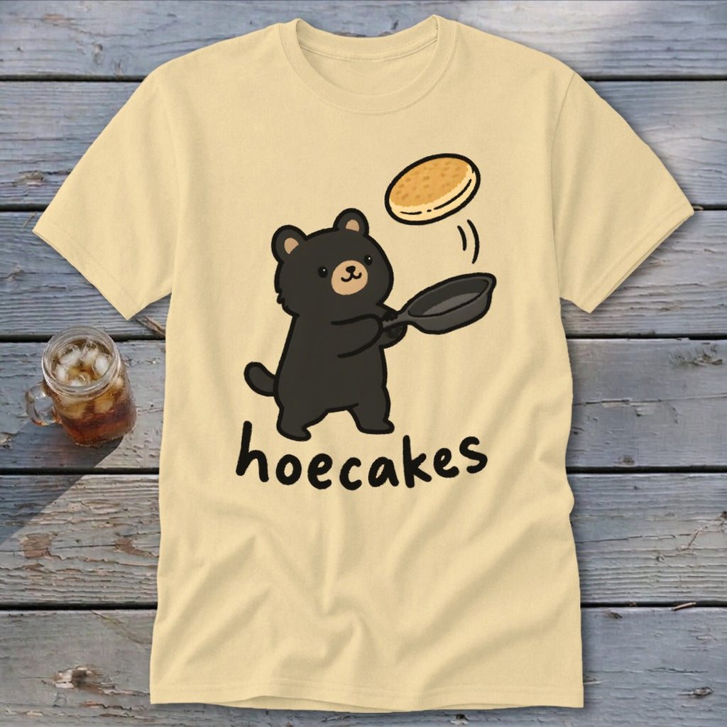 A cream-colored t-shirt featuring a cartoon black bear flipping a pancake from a skillet with the text hoecakes underneath.