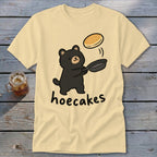 A cream-colored t-shirt featuring a cartoon black bear flipping a pancake from a skillet with the text hoecakes underneath.