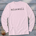 Pink long-sleeve t-shirt with black text "mizewell" on a wooden background.
