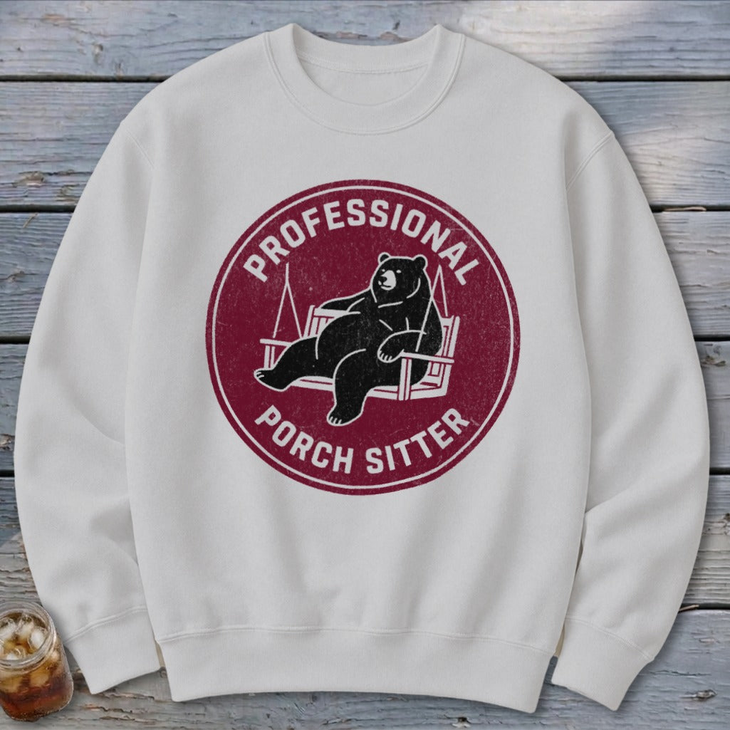 A white crewneck sweatshirt featuring a maroon circle graphic of a relaxed bear on a porch swing and the text "PROFESSIONAL PORCH SITTER".