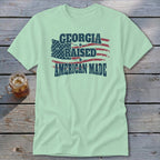 A light green t-shirt with a distressed graphic reading Georgia Raised American Made over a US flag design, lying on a wooden surface with a glass of iced tea.
