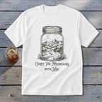 A white t-shirt featuring a mason jar illustration filled with a mountain landscape and text reading Carry The Mountains With You.