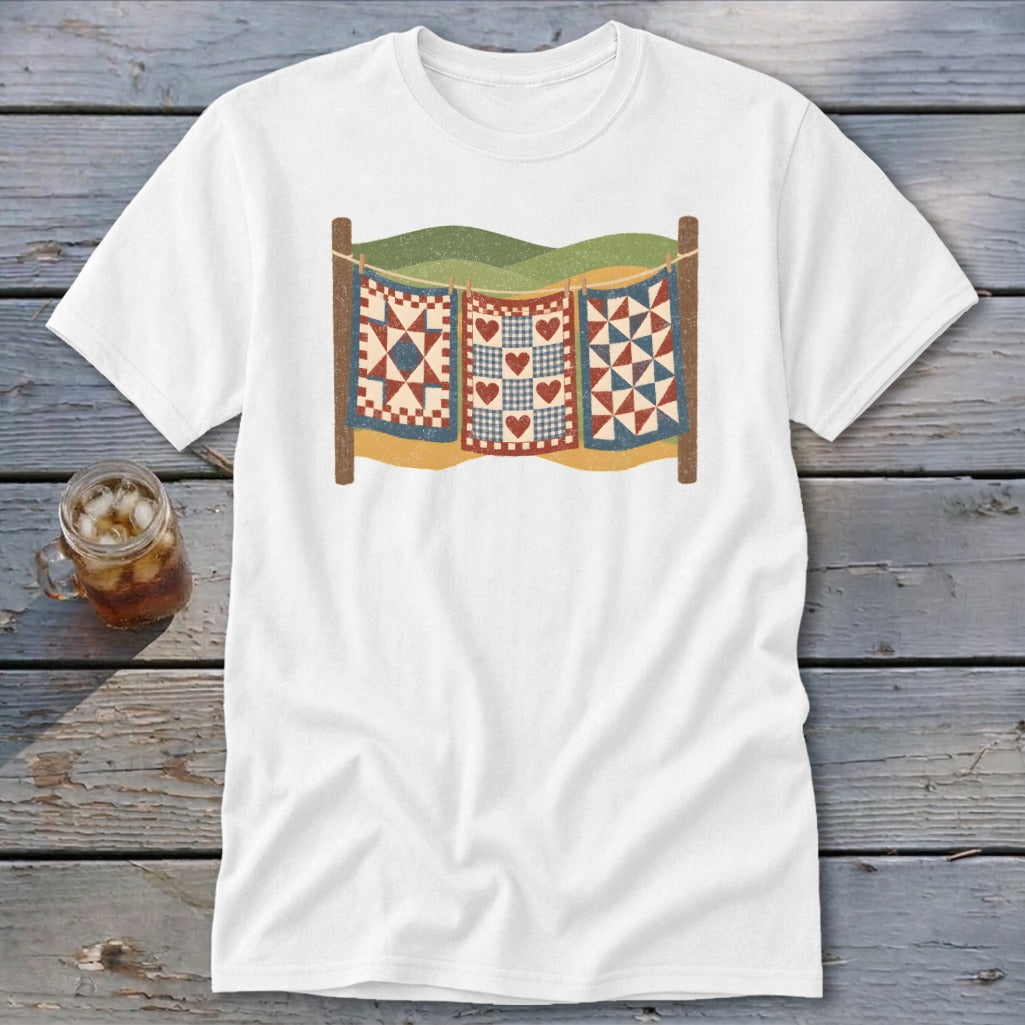A white t-shirt featuring a graphic of three patchwork quilts hanging on a clothesline against a landscape background.