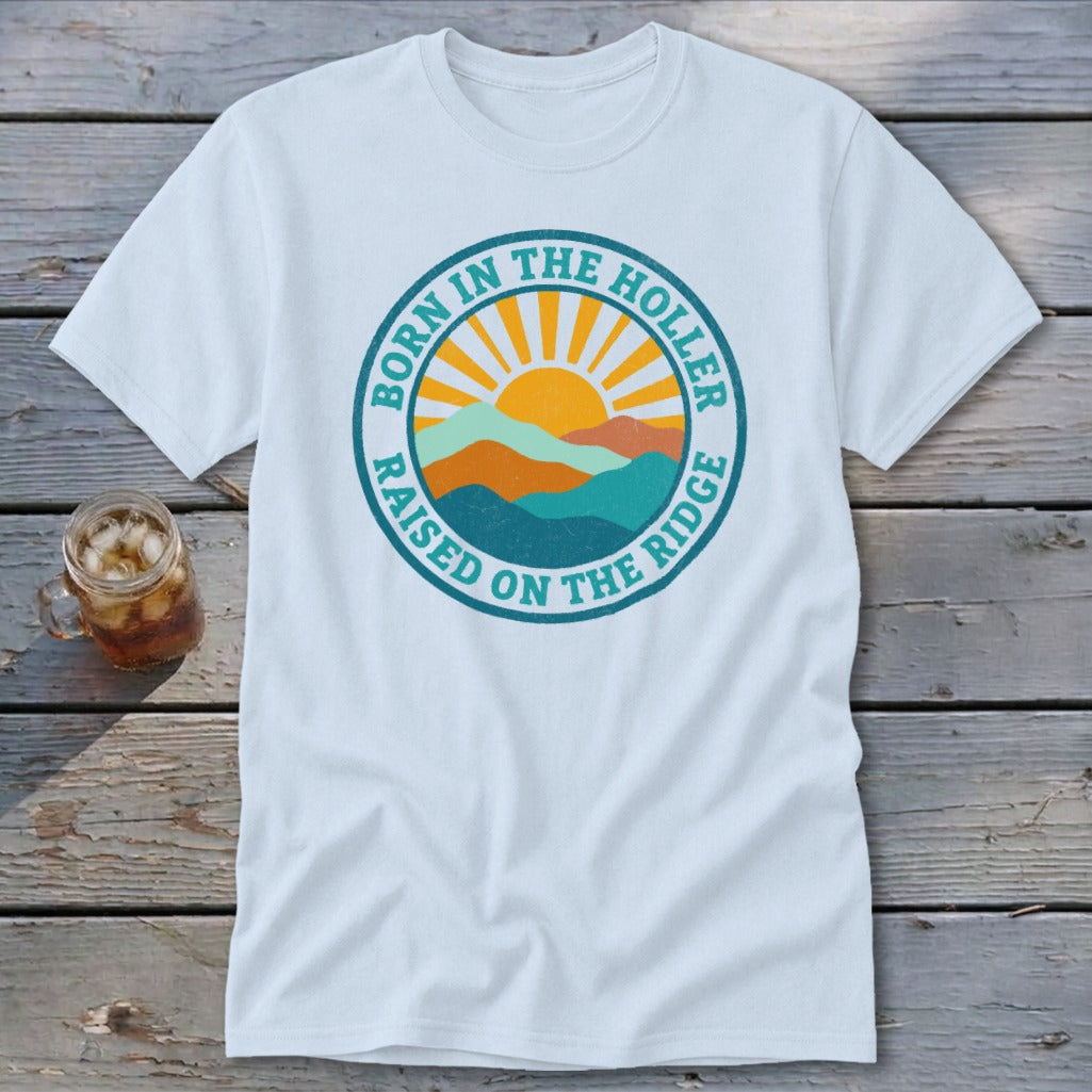 A white t-shirt featuring a retro mountain and sun graphic with text that reads Born in the Holler, Raised on the Ridge.