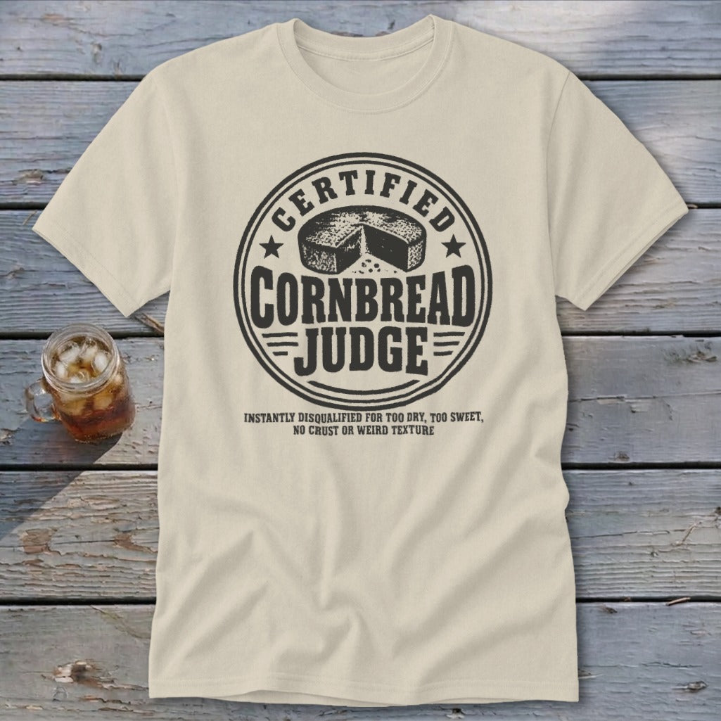 A cream-colored t-shirt featuring a black circular graphic that reads "Certified Cornbread Judge."
