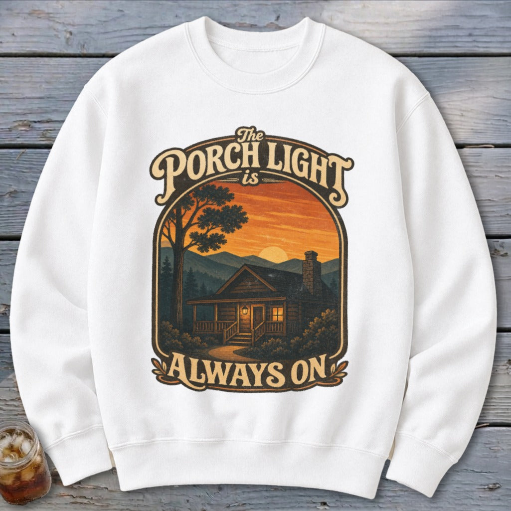 White sweatshirt with graphic: "The Porch Light is Always On" above a log cabin at sunset.