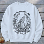 A white crewneck sweatshirt featuring a black graphic of a bird on a post surrounded by cornstalks and the text "When the whippoorwill calls it's time to plant corn".