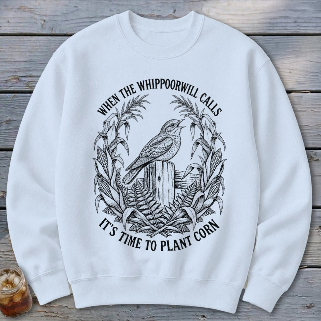 A white crewneck sweatshirt featuring a black graphic of a bird on a post surrounded by cornstalks and the text "When the whippoorwill calls it's time to plant corn".