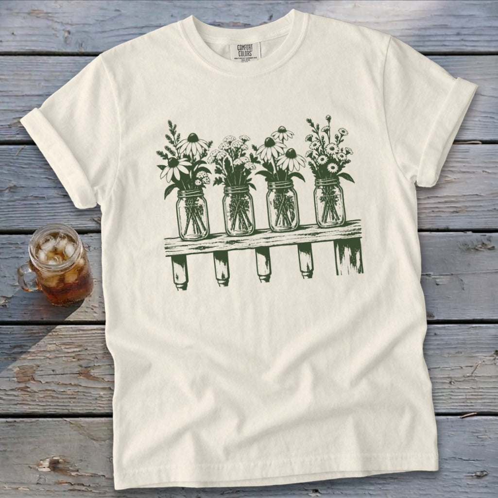 An off-white t-shirt featuring a green graphic of four mason jars filled with wildflowers on a shelf.
