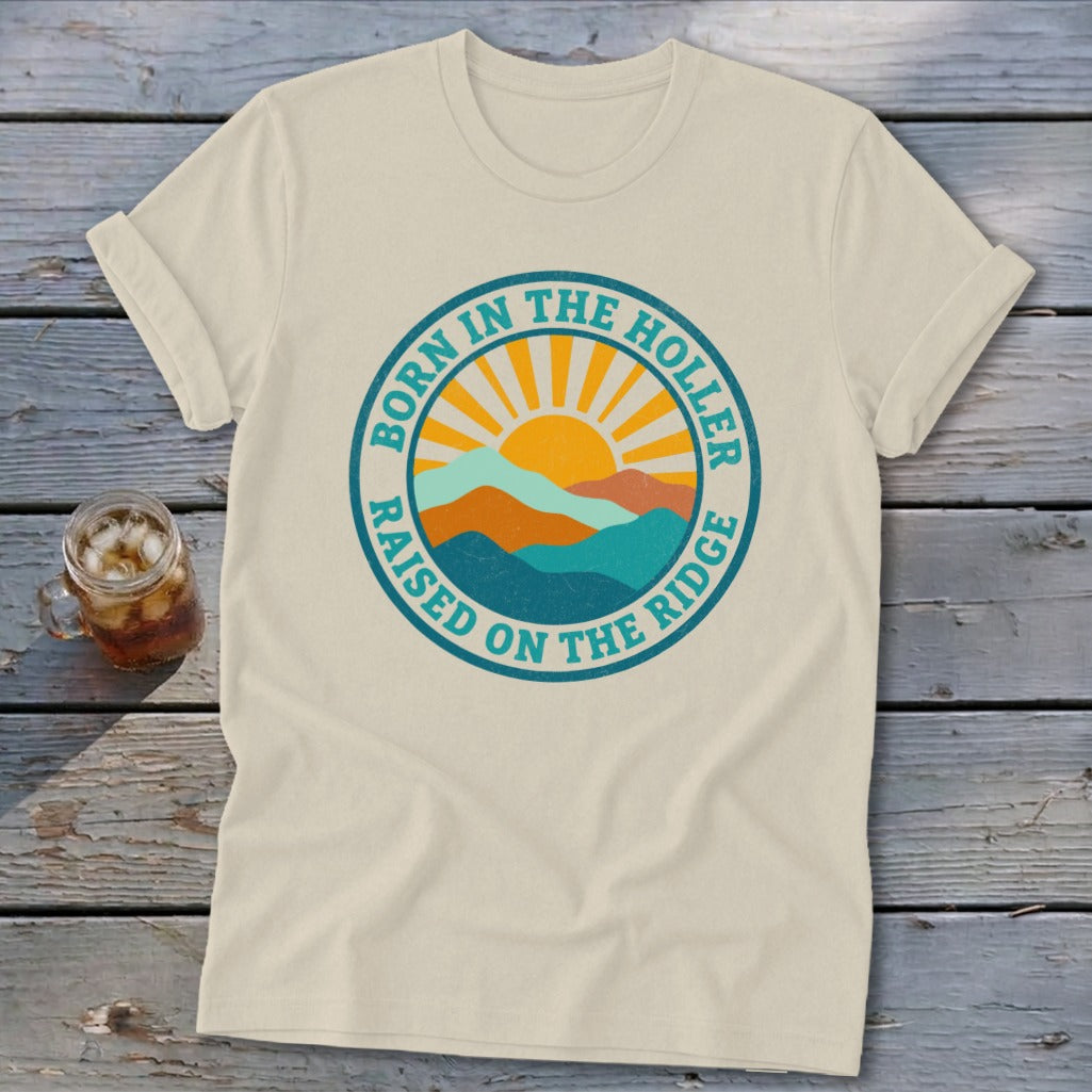 A cream-colored t-shirt with rolled sleeves features a circular retro mountain and sun graphic with text.