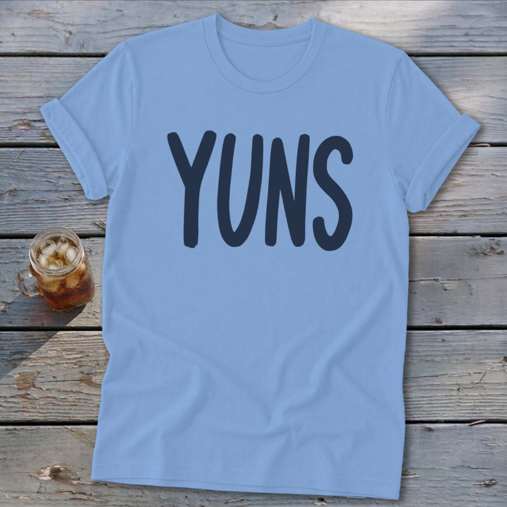 Light blue t-shirt with "YUNS" printed in navy blue, next to a glass of iced tea.