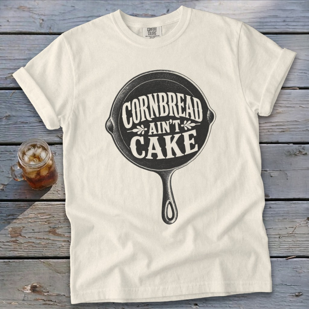 Cream t-shirt with "Cornbread Ain't Cake" slogan inside a cast iron skillet graphic.