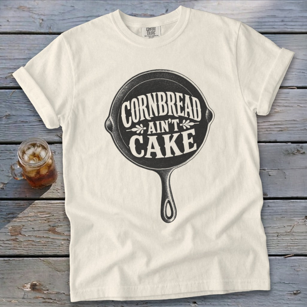 Cream t-shirt with "Cornbread Ain't Cake" slogan inside a cast iron skillet graphic.