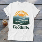 White t-shirt with a mountain and sun graphic and "The Foothills" text.