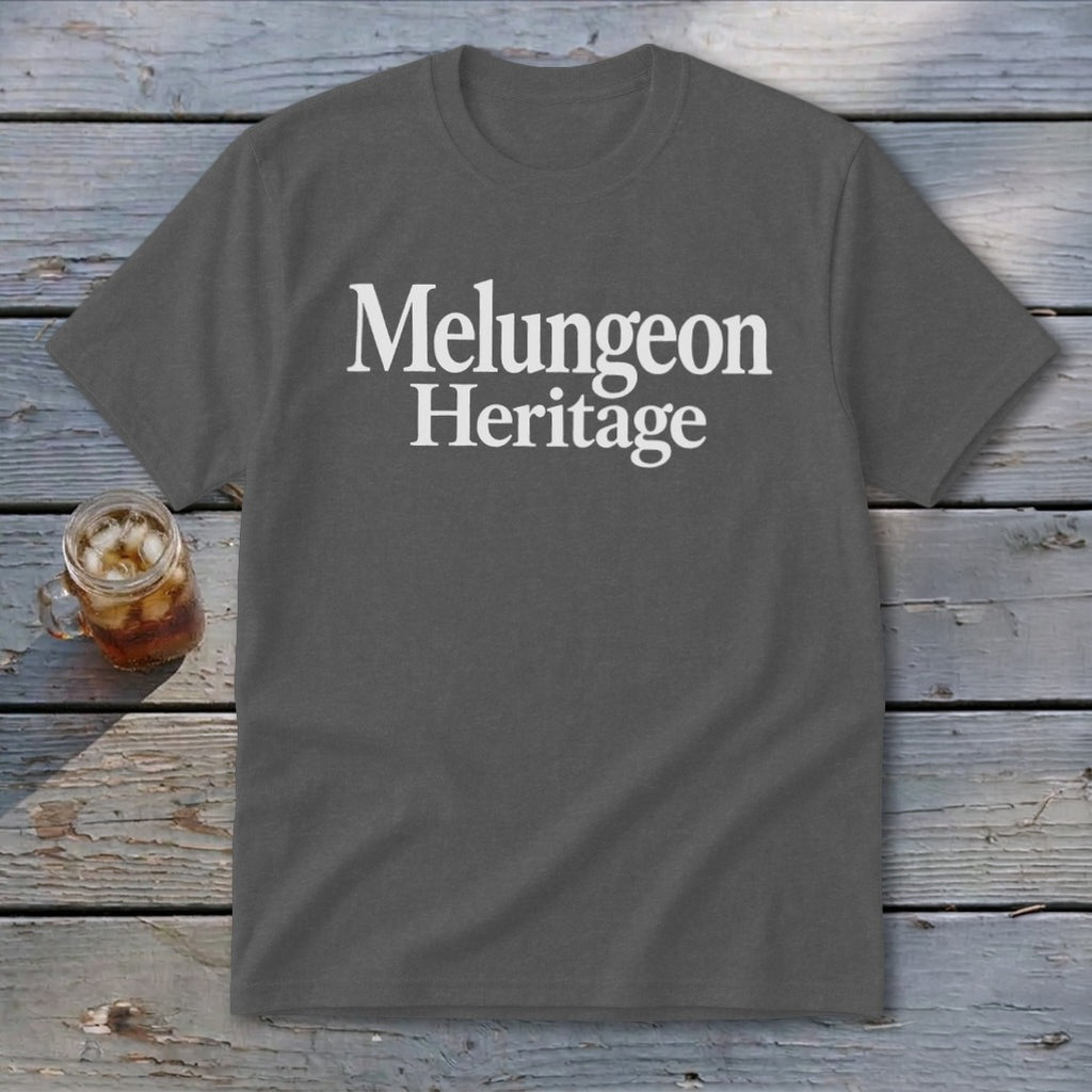 A dark gray t-shirt with the white text Melungeon Heritage centered on the chest rests on a rustic wooden background next to a glass of iced tea.