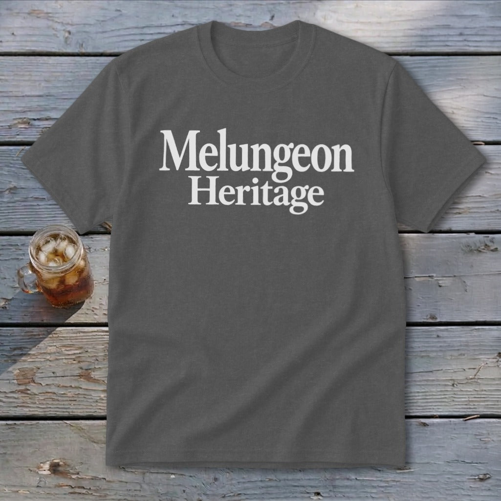A dark gray t-shirt with the white text Melungeon Heritage centered on the chest rests on a rustic wooden background next to a glass of iced tea.