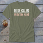 Olive green t-shirt with white and pink text reading "THESE HOLLERS KNOW MY NAME".
