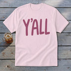 Pink t-shirt with large maroon "Y'ALL" text, set on a wooden surface.