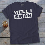 A dark blue t-shirt with white text that reads WELL I SWAN lies on a wooden surface.
