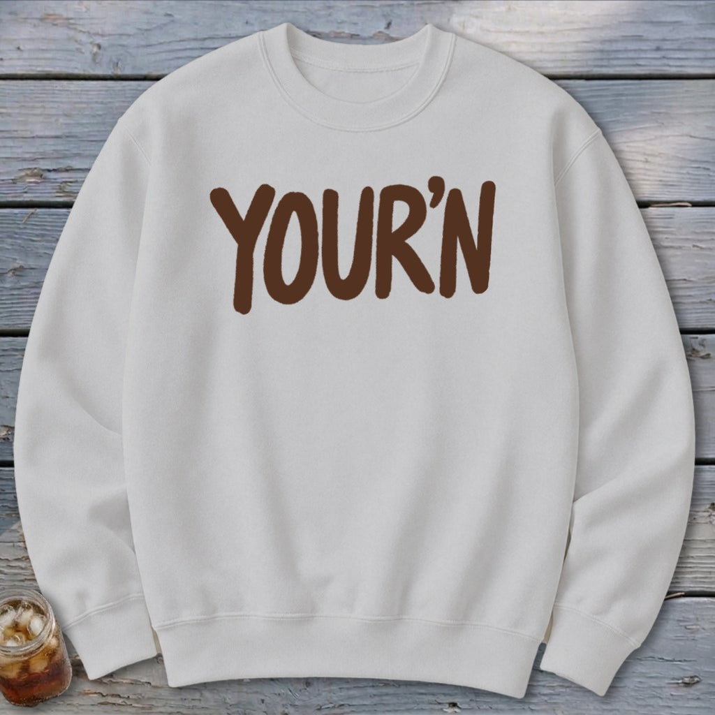 White crewneck sweatshirt with brown "YOUR'N" text, on a wooden background.