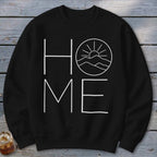 A black crewneck sweatshirt features white text spelling HOME, with a mountain and sun graphic inside the O.
