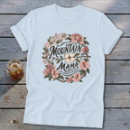 Light blue t-shirt with "Mountain Mama Raised Em Right" text within a floral wreath.