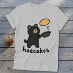 Light gray t-shirt featuring a cartoon black bear flipping a pancake in a skillet, with the text hoecakes below.
