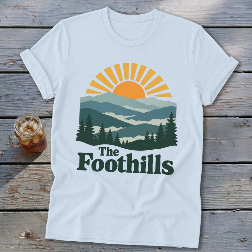 White t-shirt with mountain sunset graphic and "The Foothills" text.