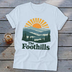 White t-shirt with mountain sunset graphic and "The Foothills" text.