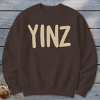 Brown crewneck sweatshirt with beige "YINZ" text, partially shown drink.