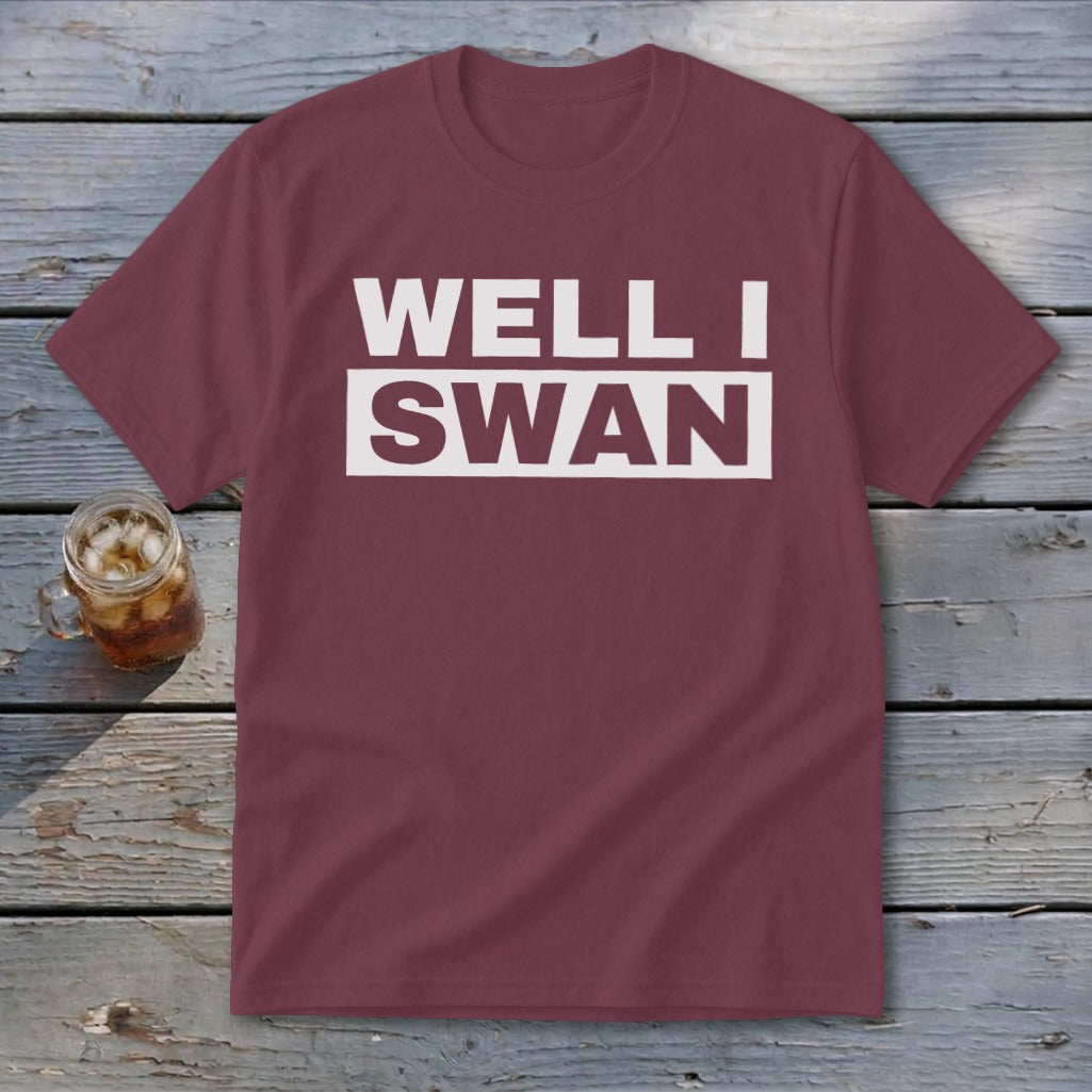 Maroon short-sleeved t-shirt with white text reading WELL I SWAN on a wooden background.