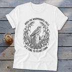 A white t-shirt featuring a black graphic of a bird on a wooden post, surrounded by corn stalks and ferns, with the text WHEN THE WHIPPOORWILL CALLS IT'S TIME TO PLANT CORN.