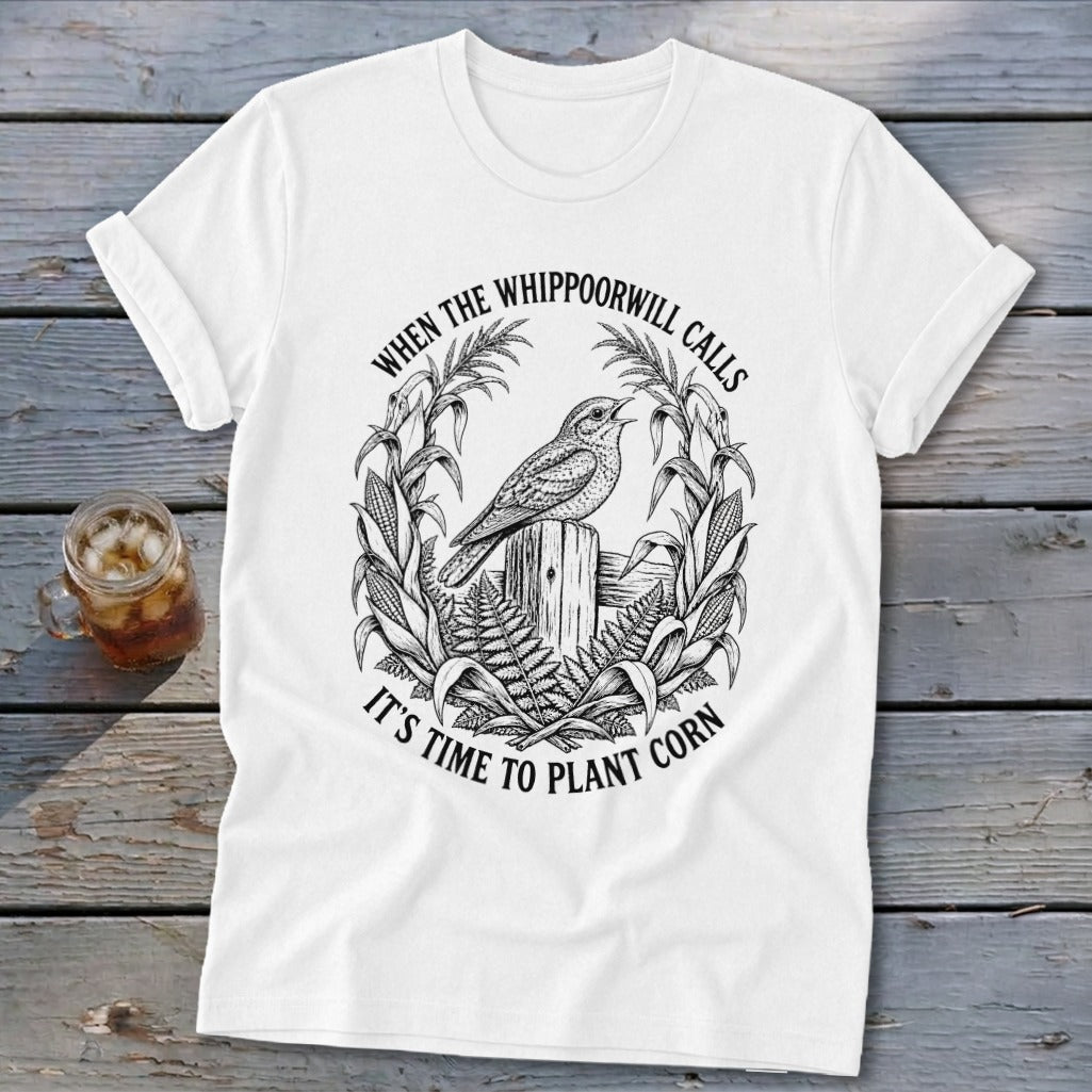 A white t-shirt featuring a black graphic of a bird on a wooden post, surrounded by corn stalks and ferns, with the text WHEN THE WHIPPOORWILL CALLS IT'S TIME TO PLANT CORN.