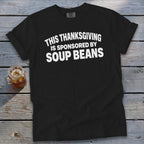 Black t-shirt with white text reading THIS THANKSGIVING IS SPONSORED BY SOUP BEANS on a wooden background.