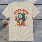 Cream t-shirt with distressed graphic of a bear wearing a cap, drinking from a jar. Text reads "Porch Sittin' Club".