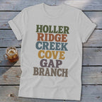 A heather gray t-shirt featuring stacked multicolored retro text resting on a rustic wooden surface.