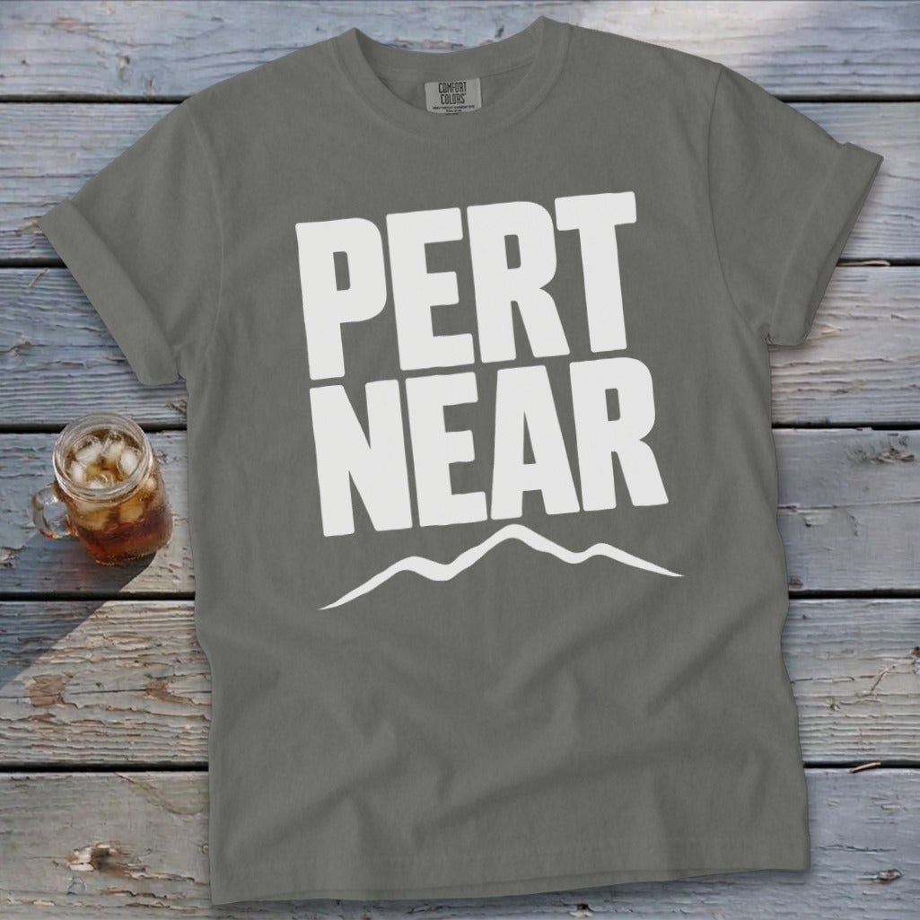 Gray crewneck t-shirt with white text reading PERT NEAR above a mountain outline, lying on a wooden surface beside a glass of iced tea.