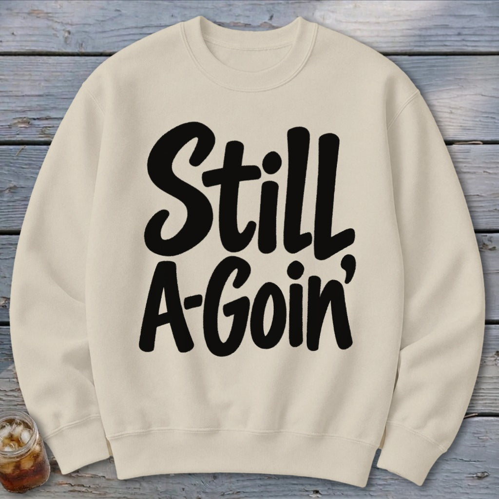 A cream-colored crewneck sweatshirt featuring the black graphic text Still A-Goin' laid flat on a rustic wooden surface.