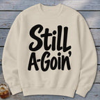 A cream-colored crewneck sweatshirt featuring the black graphic text Still A-Goin' laid flat on a rustic wooden surface.