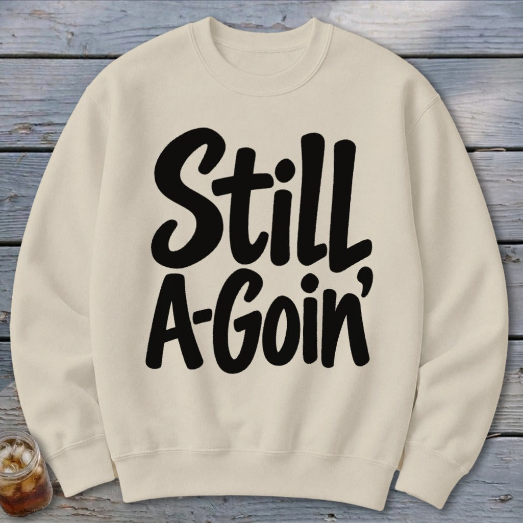 A cream-colored crewneck sweatshirt featuring the black graphic text Still A-Goin' laid flat on a rustic wooden surface.
