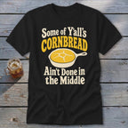 A black t-shirt printed with yellow and white text reading "Some of Y'all's Cornbread Ain't Done in the Middle" and a skillet of cornbread.