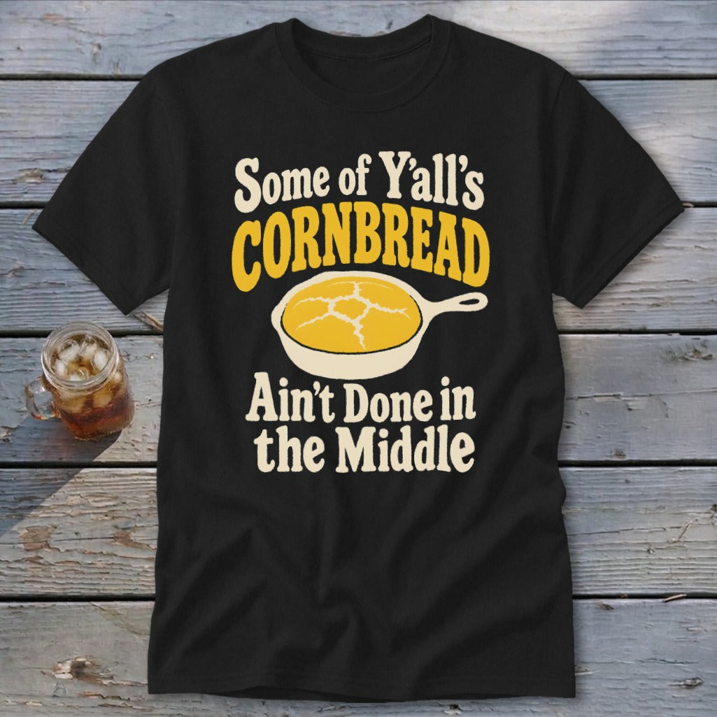 A black t-shirt printed with yellow and white text reading "Some of Y'all's Cornbread Ain't Done in the Middle" and a skillet of cornbread.