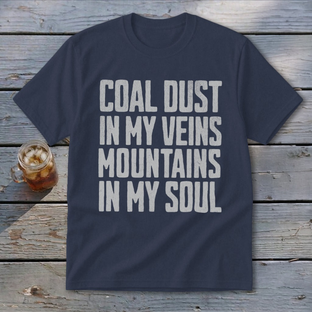 A navy blue t-shirt displaying the text COAL DUST IN MY VEINS MOUNTAINS IN MY SOUL in white, lying on a wooden surface with a glass of iced tea.