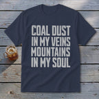 A navy blue t-shirt displaying the text COAL DUST IN MY VEINS MOUNTAINS IN MY SOUL in white, lying on a wooden surface with a glass of iced tea.