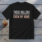 Black t-shirt with white text "THESE HOLLERS" and rose gold text "KNOW MY NAME".
