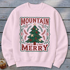 A light pink crewneck sweatshirt featuring a graphic of a Christmas tree with red berries, framed by red text reading MOUNTAIN MERRY.