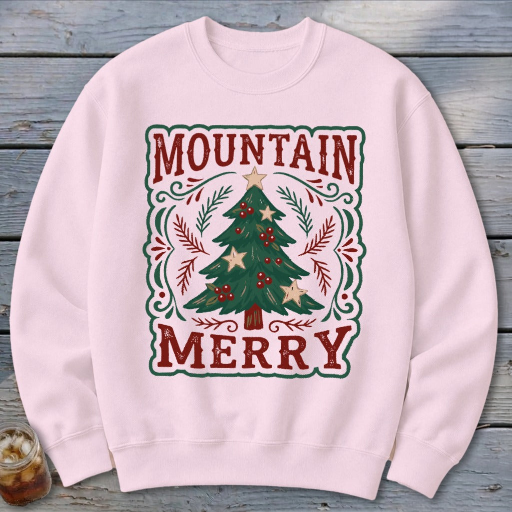 A light pink crewneck sweatshirt featuring a graphic of a Christmas tree with red berries, framed by red text reading MOUNTAIN MERRY.
